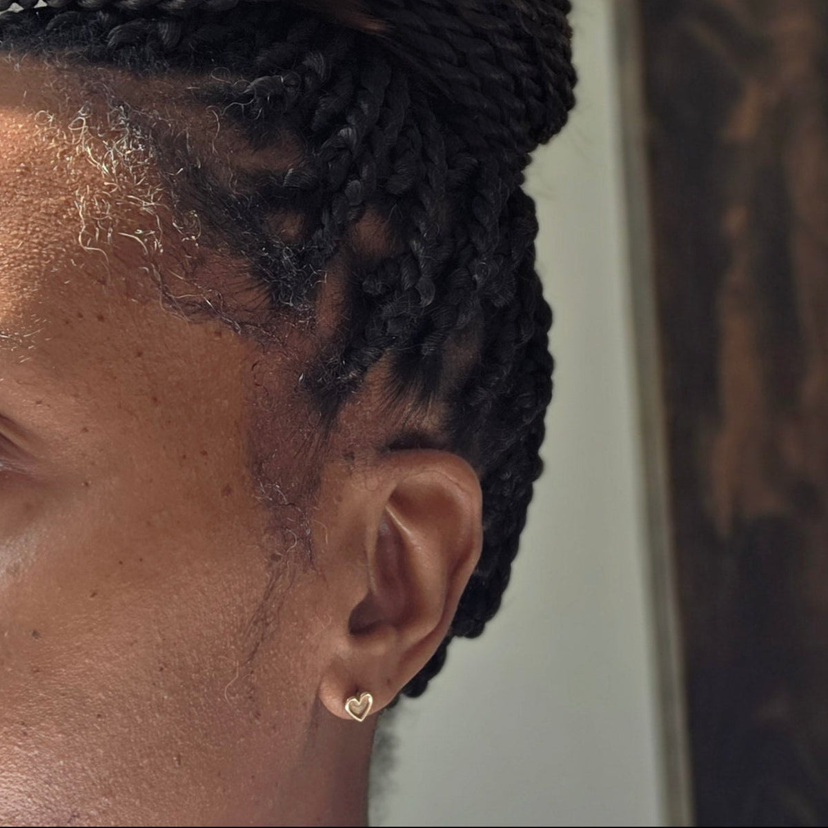 A close-up side profile of a person with braided black hair and a small 9ct gold heart-shaped stud earring in their earlobe.
