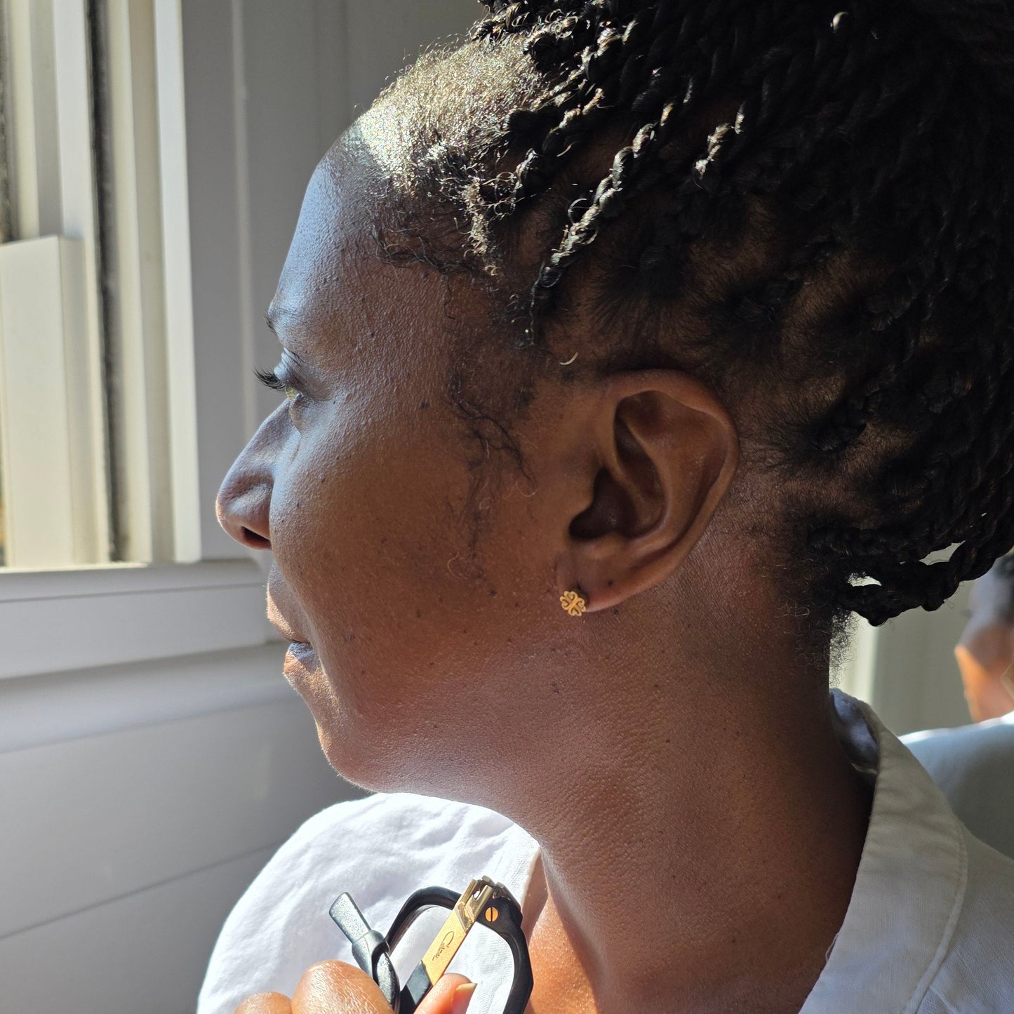 A close-up side profile of a woman with braided hair wearing a small gold stud earring, called Nyame Dua photographed indoors near a window.