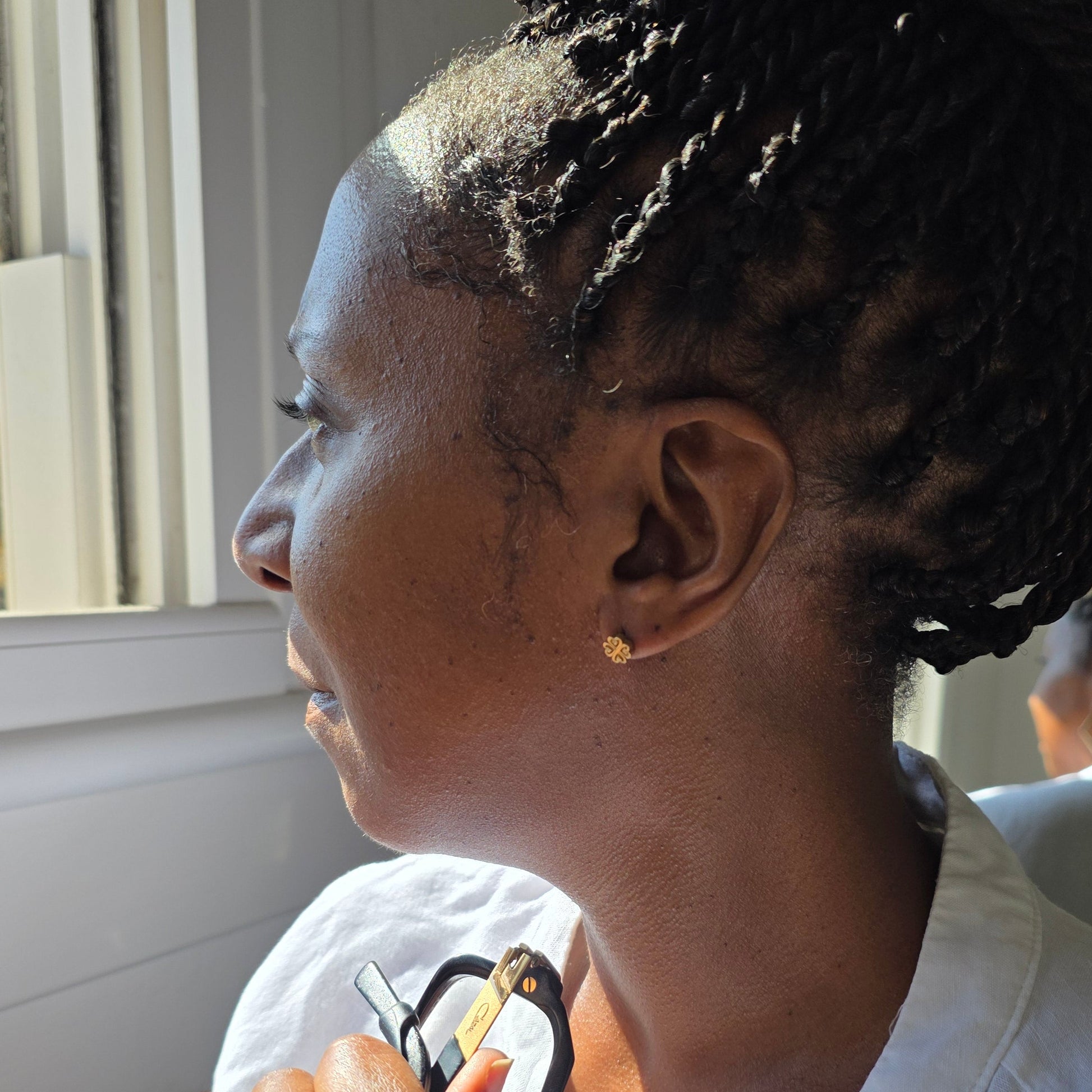 A close-up side profile of a woman with braided hair wearing a small gold stud earring, called Nyame Dua photographed indoors near a window.